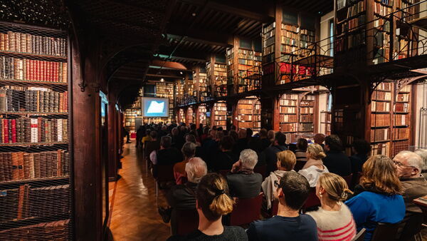 Een zittend publiek luistert naar een lezing in de historische Nottebohmzaal, omringd door houten boekenkasten vol oude boeken.
